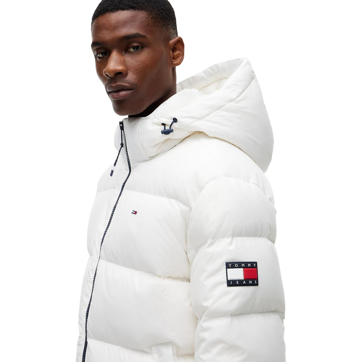 Piumino bianco uomo Tommy Jeans DM0DM19786YBL. Giambotto imbottito in piuma d'anatra relaxed fit oversize con cappuccio, zip integrale e logo Tommy ricamato.