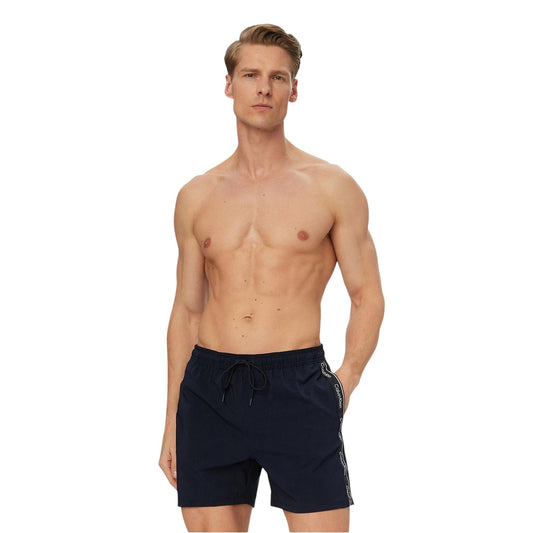 Boxer Mare CK Jeans Blu; Bande Laterali Logate. 10% Elastam per massima elasticità, poliestere riciclato, asciugatura rapida. Tasche laterali, vita con coulisse, fit regolare e sportivo Vista frontale indossato da modello 