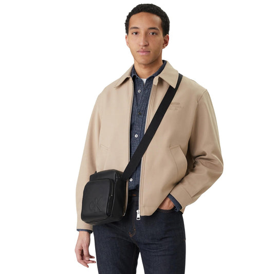 Borsa reporter Calvin Klein in ecopelle; monogramma stampato, tasche con zip, tracolla regolabile. Accessorio compatto ed elegante. Colore nero indossato da modello 